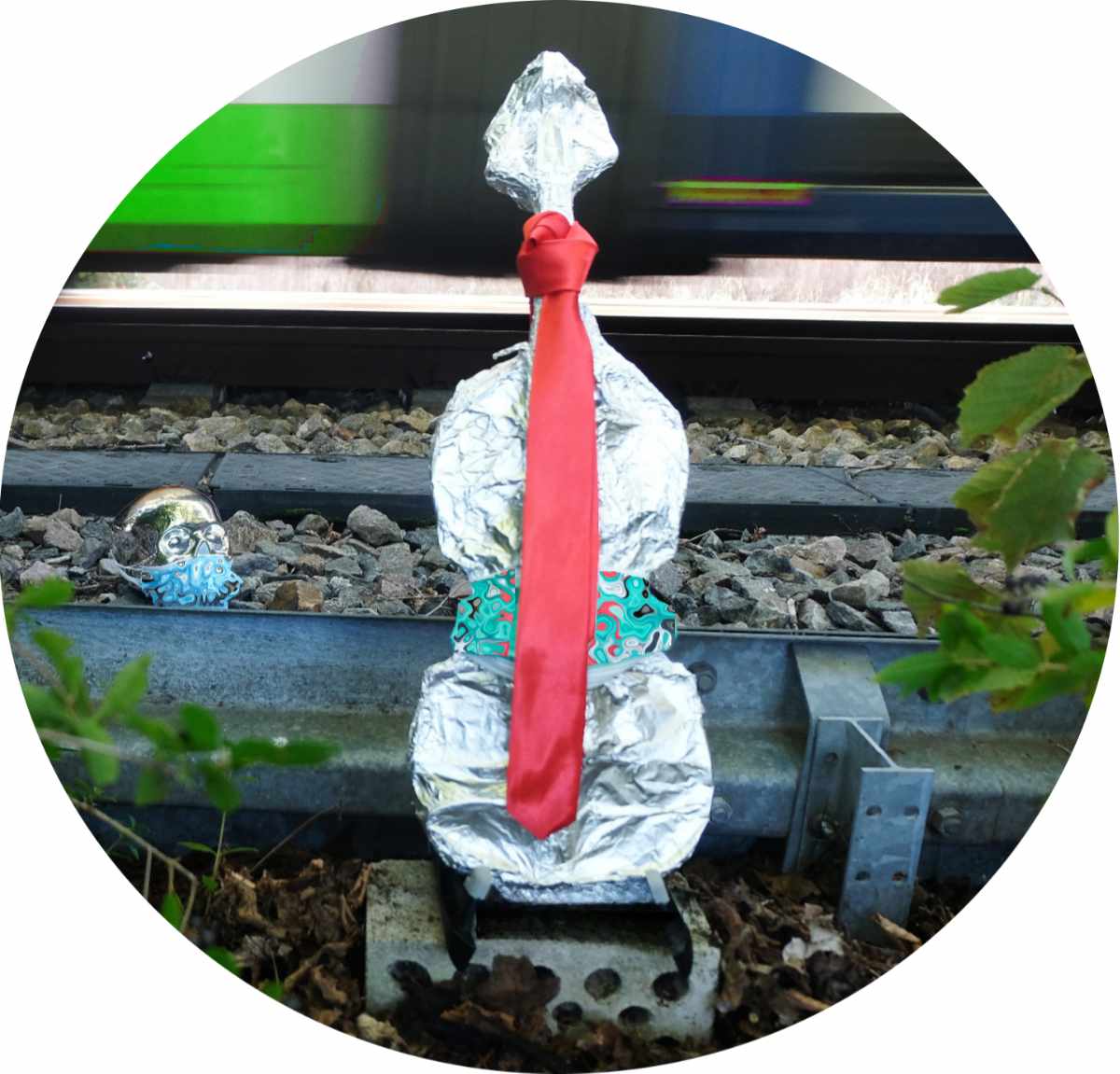 Eine Violine ist in Alufolie verpackt, trägt rote Lederkrawatte, steht an einem Bahngleis, ein Zug rast durch. Dahinter ein Totenkopf als Sparbüchse mit ffp2-Maske.