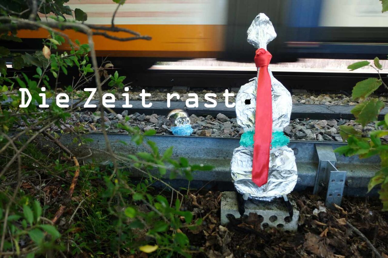 Geige in Alufolie verpackt und zwischen zwei Städten vor Zuggleise gestellt, ein Zug fährt gerade durch.