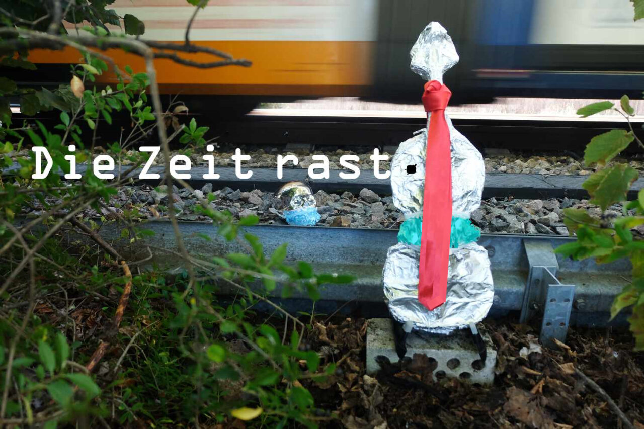Eine Geige ist in Alufolie verpackt. Sie wird an einem Zuggleis abgestellt, an dem ein Zug durchrast. Symbolisch dafür: "Die Zeit rast." Mit schwarzem, quadratischem Punkt auf der silberenen Alufolie der Geige, auf dieser links oben. Die Geige trägt eine rote Lederkrawatte und eine verschwommen gestaltete ffp2-Maske.